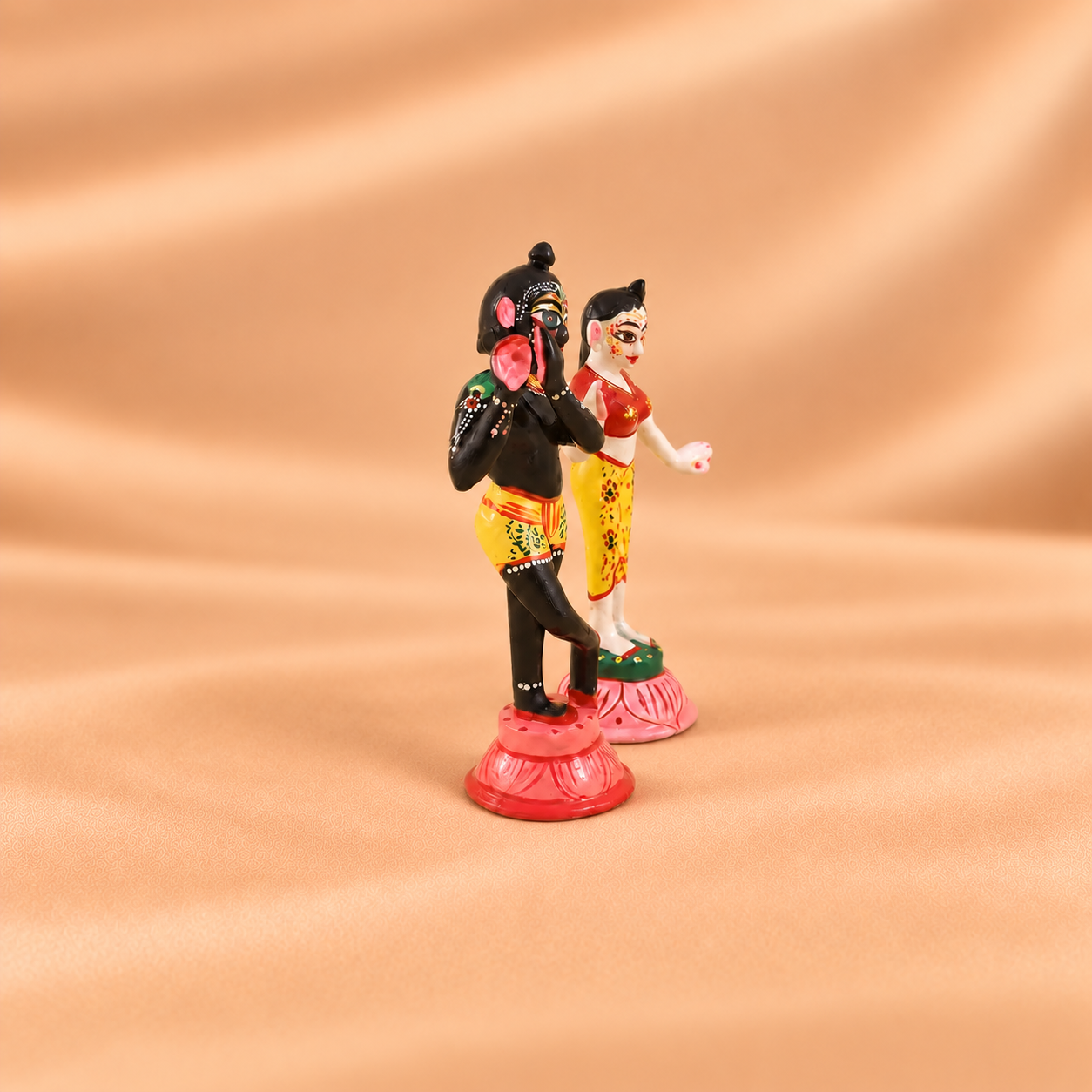 A pair of traditional figurines depicting Lord Krishna and a woman, standing on pink lotus bases against a draped beige fabric background.