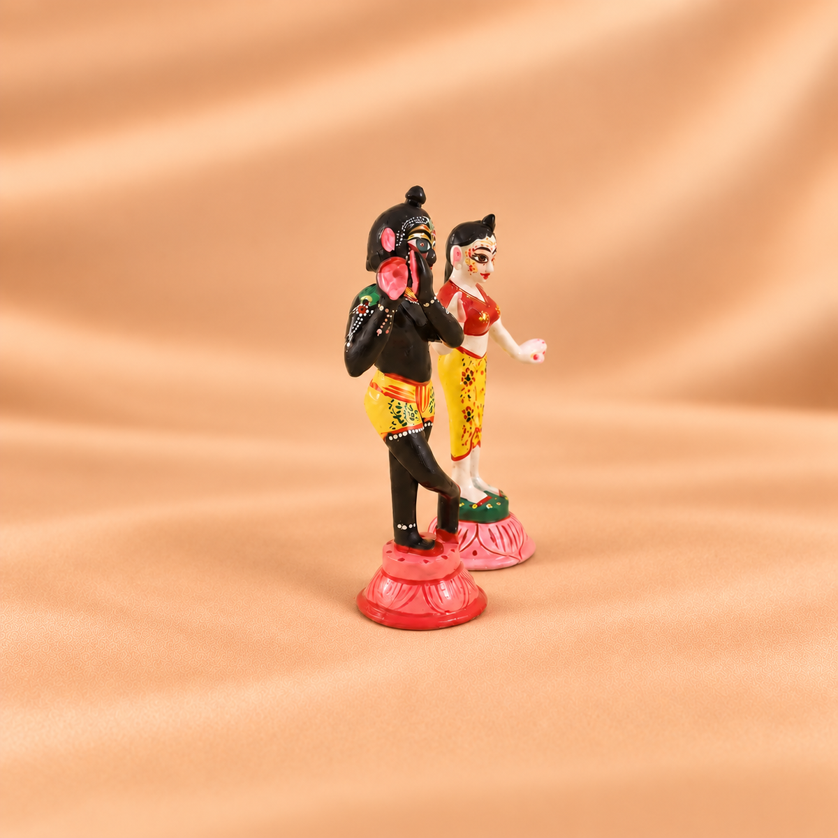 A pair of traditional figurines depicting Lord Krishna and a woman, standing on pink lotus bases against a draped beige fabric background.