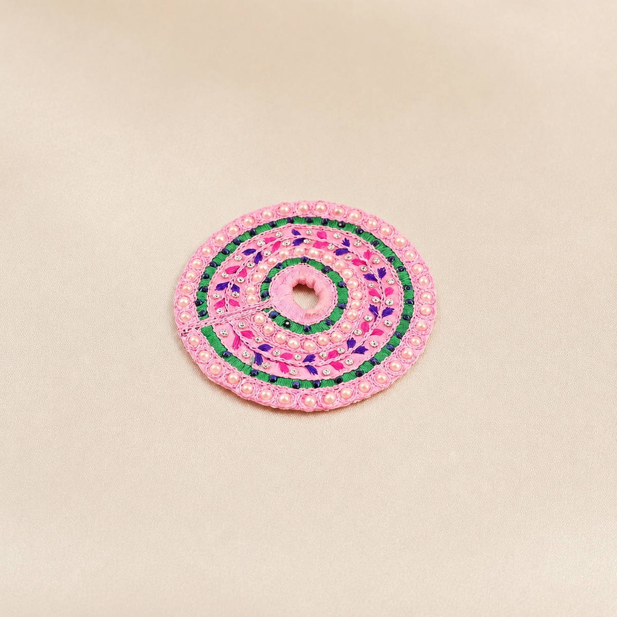 Round pink Laddu Gopal dress with pearl border, green and purple threadwork, rhinestones and floral embroidery on an ivory satin background.