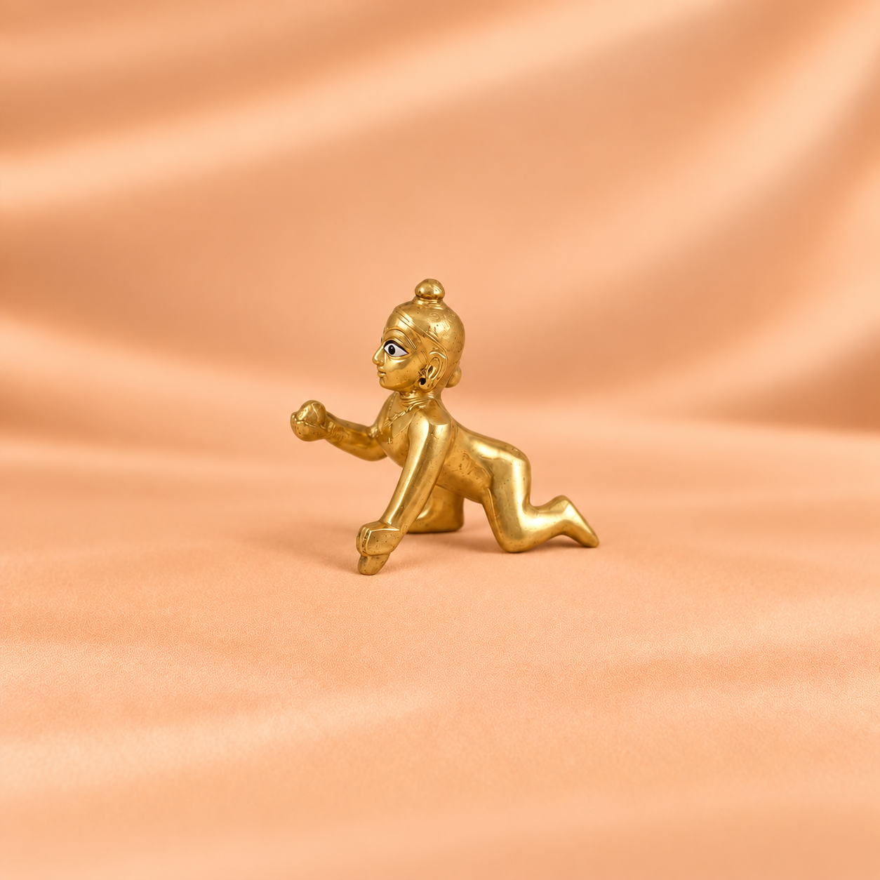 Side view of a plain golden crawling Laddu Gopal idol holding a laddu against a peach background.