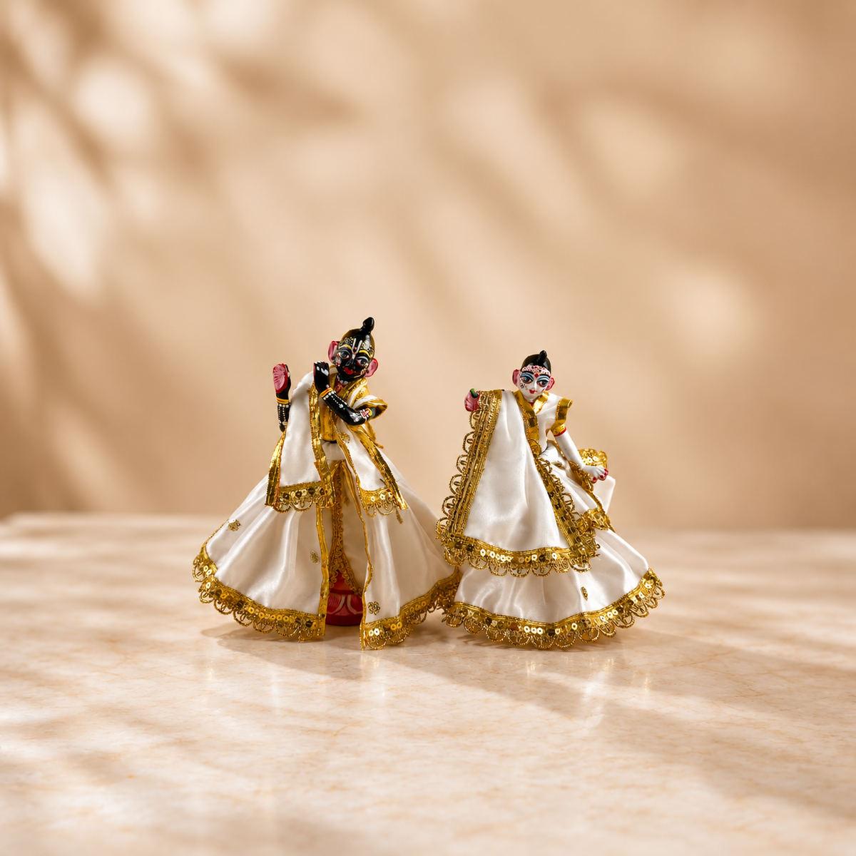 Radha Krishna idols wearing white poshak with ornate gold-tone lace and sequins on a cream marble surface against a warm beige background.