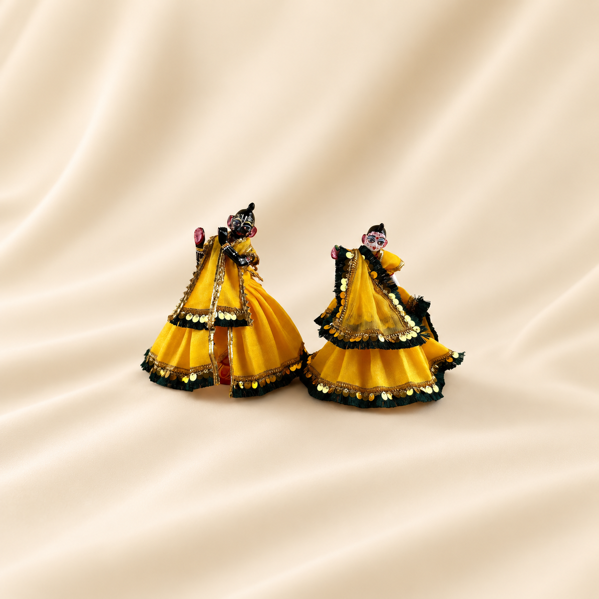 Radha Krishna idols wearing yellow poshak with dark fringe and gold-tone sequin trim, placed together on cream fabric.