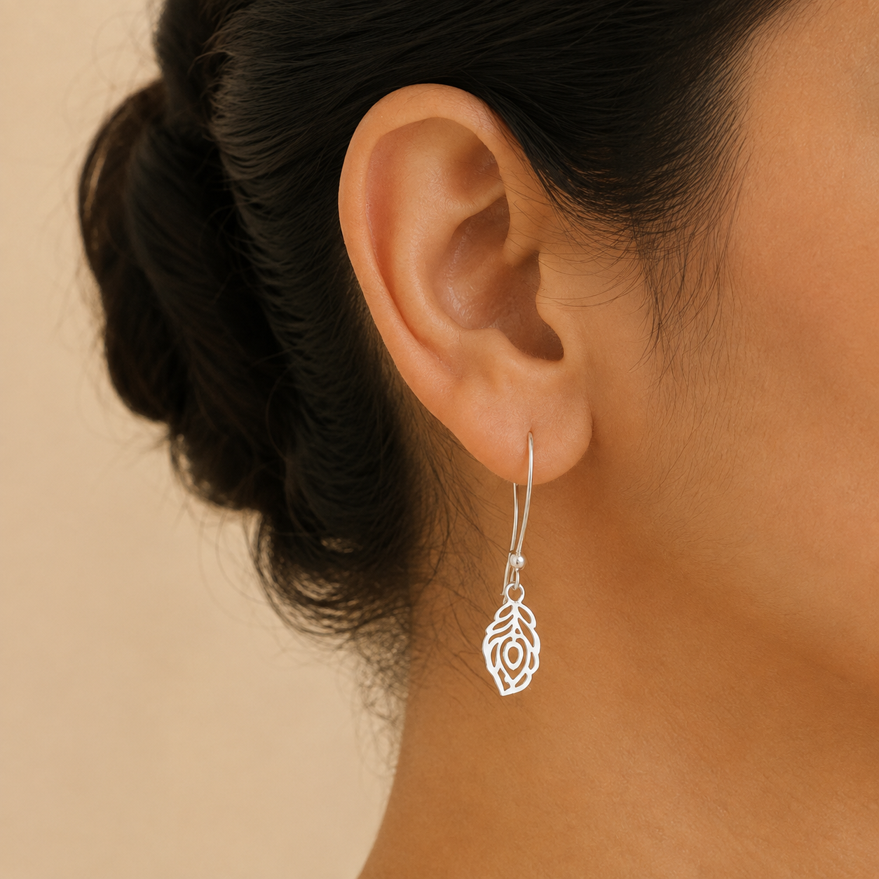 A close-up of a person's ear wearing a delicate silver dangle earring with an intricate cutout leaf design, set against a soft beige background.