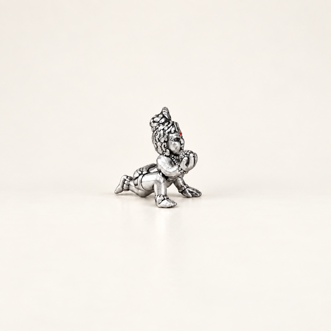 Side view of a silver crawling Laddu Gopal idol holding a sweet against an off-white background.