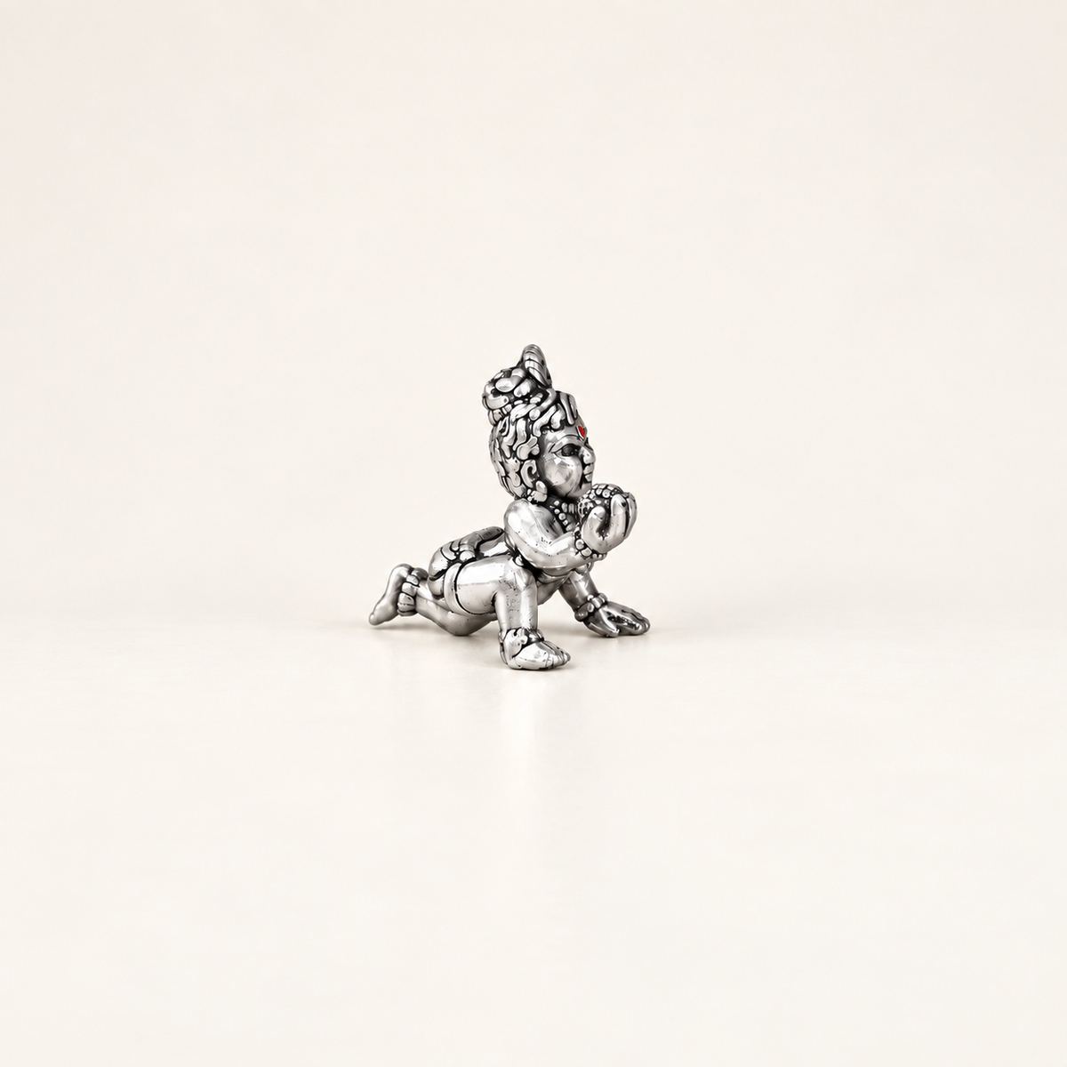 Side view of a silver crawling Laddu Gopal idol holding a sweet against an off-white background.