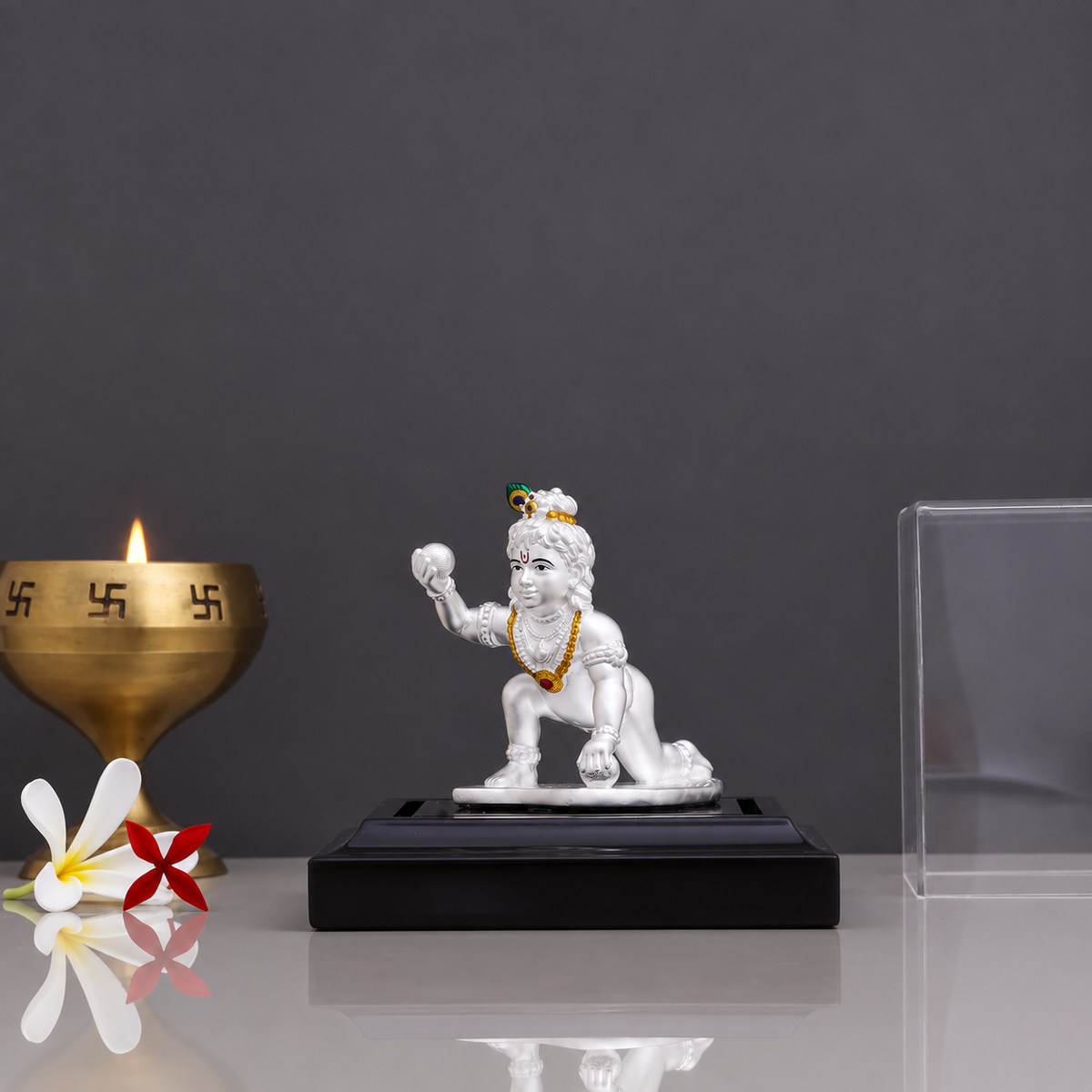 Silver Laddu Gopal idol on a black display stand with a lit brass diya.