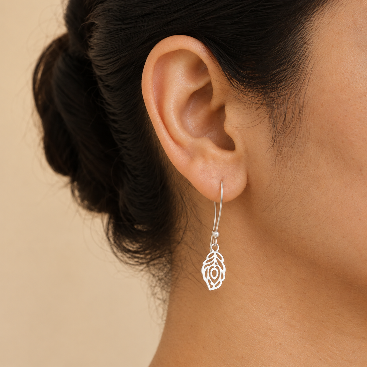 Silver peacock feather cutout earring with a long hook worn on the ear.