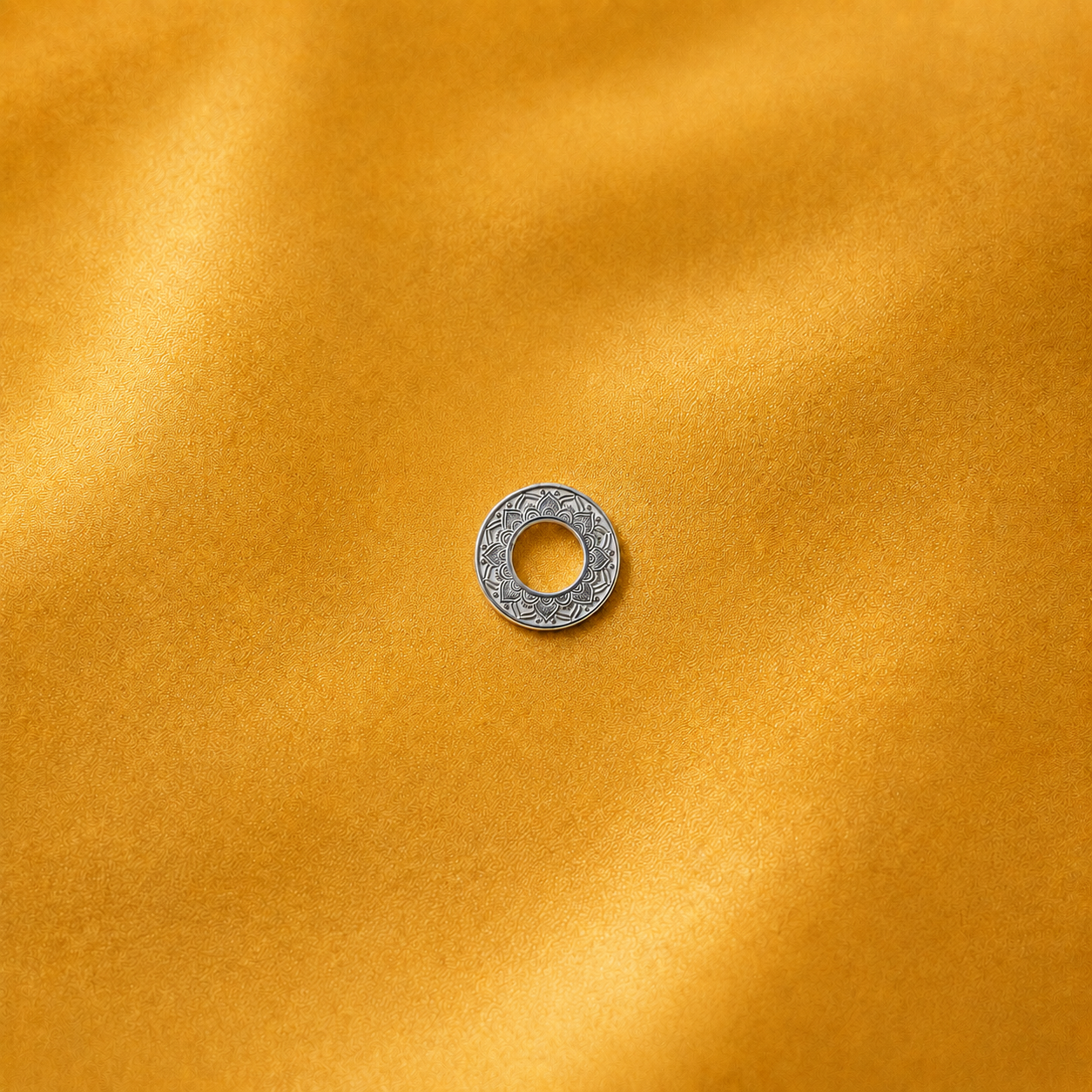 A small silver-toned circular ring charm with a textured, patterned outer edge, centered on a draped golden-yellow fabric background.