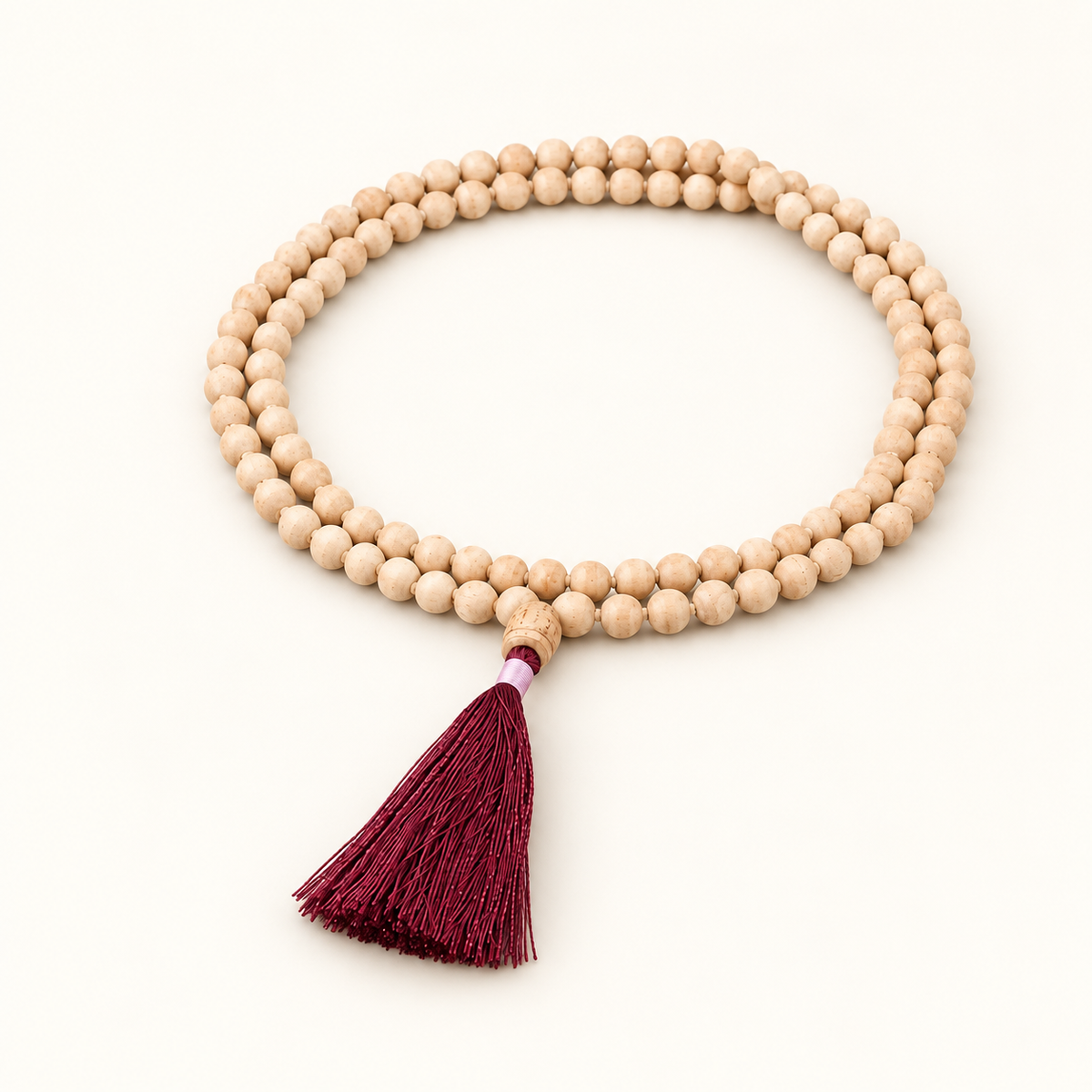 Light wooden Tulsi japa mala arranged in two loops with a burgundy tassel on an off-white background.