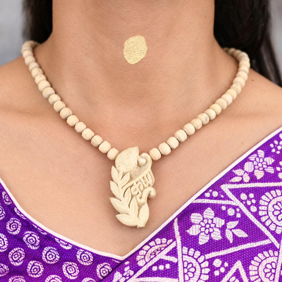 Tulsi mala with rounded wooden beads and a carved Krishna pendant with a peacock feather design, worn with a purple saree.